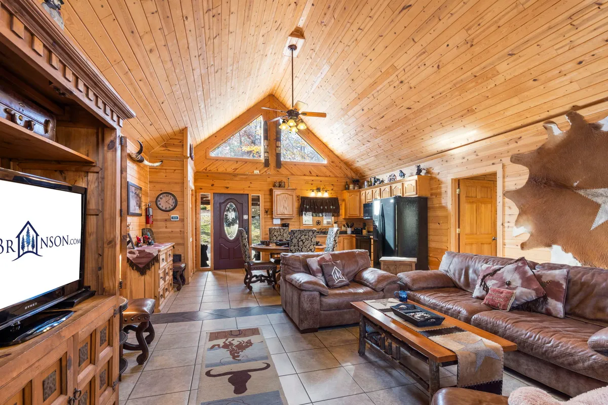 The image showcases the spacious, open-concept living area of a luxury log cabin, featuring soaring wooden ceilings, plush comfortable couches, a large dining area, and expansive windows that flood the space with natural light. The setting invites guests to relax, socialize, and enjoy cozy evenings or memorable gatherings.