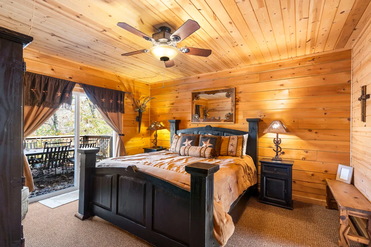 A serene bedroom with a bed adorned with decorative pillows, wood-paneled ceiling and walls, a large window offering scenic outdoor views, and nightstands with lamps. The space has a calm and peaceful atmosphere perfect for unwinding.