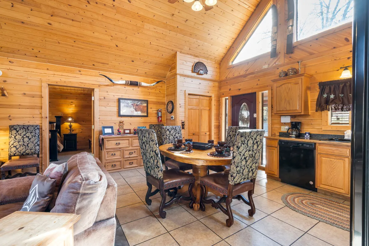 This image shows a large wooden dining table with ornate chairs, kitchen cabinetry in the background, and large windows that fill the space with natural light. The layout invites group dining, game nights, or celebration feasts in a cozy, welcoming setting.