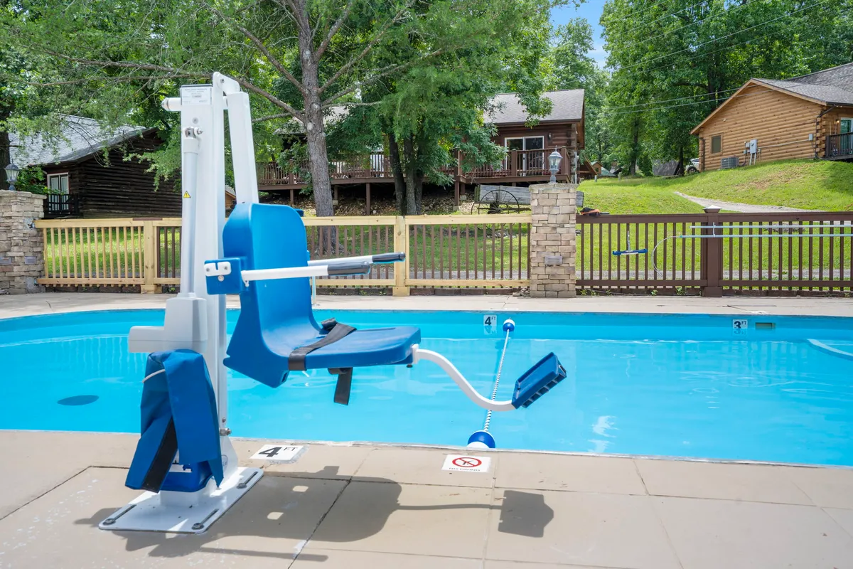 A spacious swimming pool surrounded by trees and wooden cabins, equipped with a pool lift for accessibility. Lounge chairs line the poolside, inviting guests to gather, swim, or soak up the sun in a communal, yet serene setting.