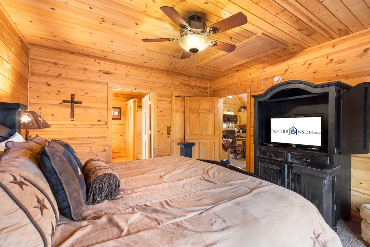 A king-sized bed with plush bedding is set against warm wooden walls. The room features a television, ceiling fan, and subtle rustic decor, creating a welcoming space for relaxation and entertainment.