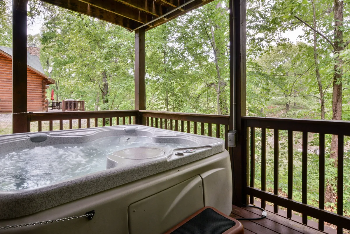 A bubbling hot tub sits on a wooden lower deck surrounded by trees, with sunset light filtering through. The inviting setup offers a tranquil spot for a soothing soak overlooking the woods.