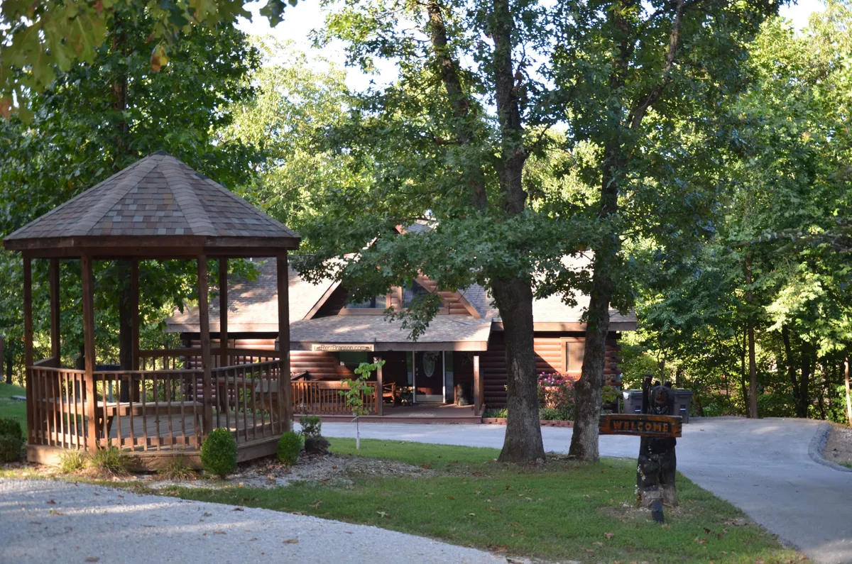 The image shows the welcoming approach to the Lone Star Cabin, including a spacious paved driveway, the main house, a charming gazebo, and a visible welcome sign. The driveway suggests ample parking, and the overall scene is tranquil and inviting.