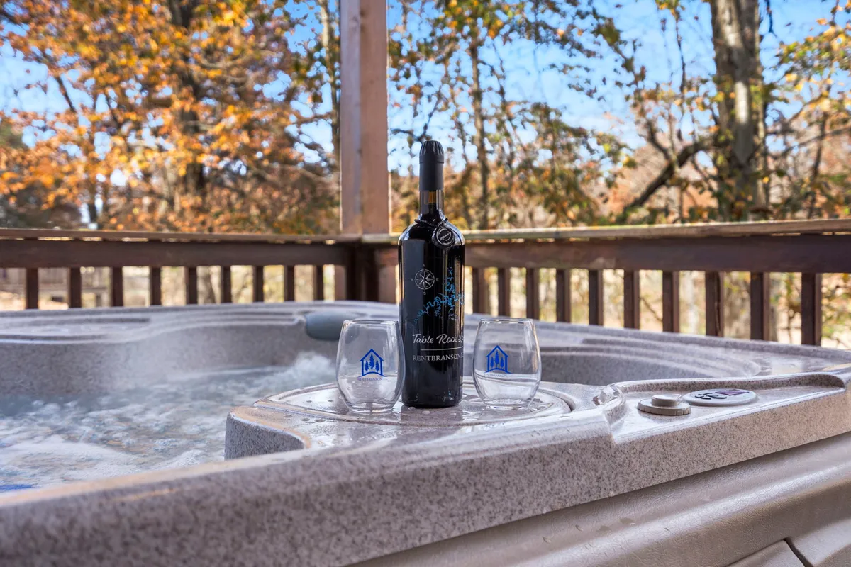 This image features an outdoor hot tub set on a wooden deck, accompanied by a wine bottle and glasses. The surrounding natural scenery is visible, creating a tranquil setting ideal for relaxation and social gatherings.