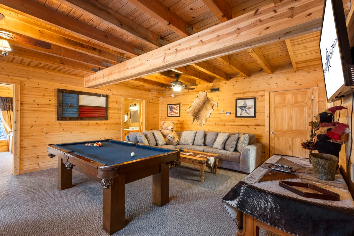 A cozy lower-level lounge featuring a pool table, large comfortable sofa, rustic wooden ceiling, and decorative wall hangings. The scene is set for fun and relaxation, with ample space for family members to play games, watch TV, or unwind together.