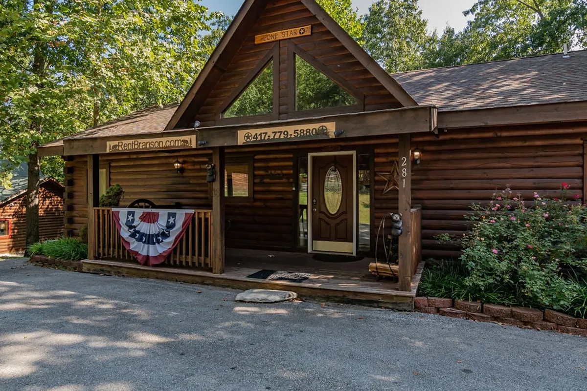 An inviting exterior shot of a log cabin with a charming front porch, decorative banner, and vibrant flower beds. The porch offers a cozy spot for enjoying the fresh Ozark air, whether for morning coffee or sunset views.