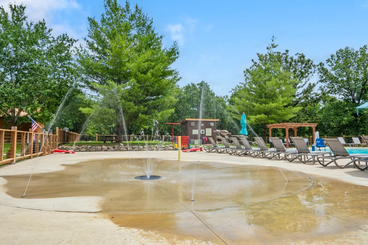 A lively exterior scene with a splash pad surrounded by lounge chairs and water features. Children can be seen playing, while families relax nearby, all bathed in bright sunlight. The joyful energy captures the essence of summer fun and outdoor activity at the resort.